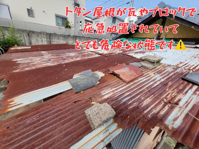 全体的に赤錆が広がったトタン屋根。瓦やブロックで応急処置がされており、雨漏りと飛散の危険がある状態。