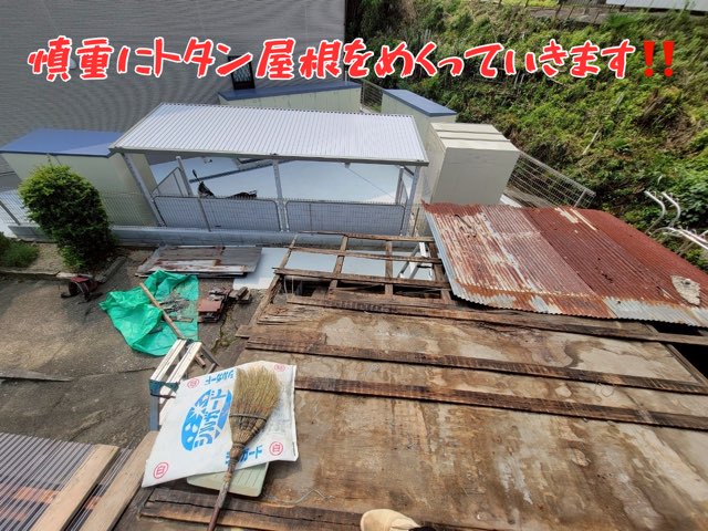 一部トタンが取り外され、下地の木材が露出している様子。地面には緑のシートや資材が広げられている。