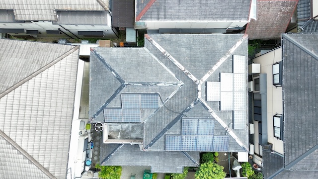 ドローンで真上から撮影したスレート屋根の全景。複雑な屋根形状に合わせて太陽光パネルが設置されているが、白い線のように見える「棟板金（むねばんきん）」の塗装が剥がれ、劣化している様子が確認できる。